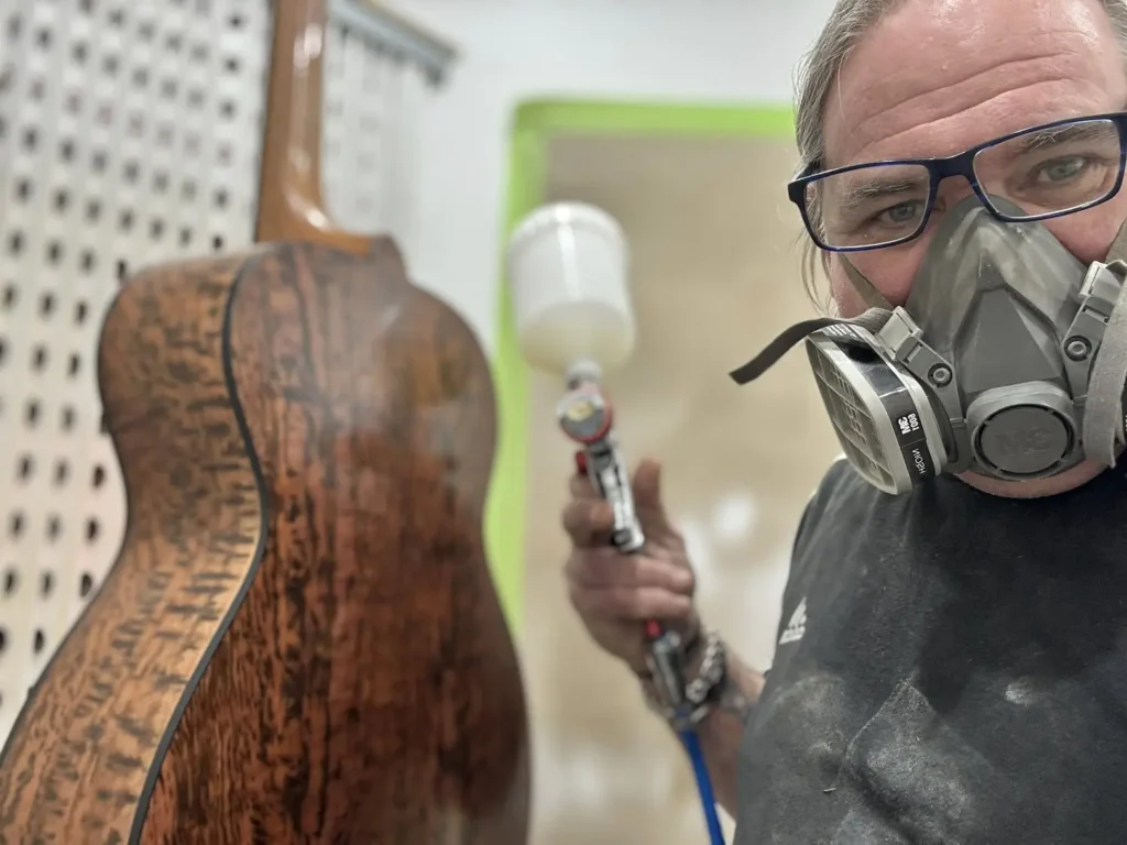 Andy in the spray booth wearing a respirator mask spraying a guitar